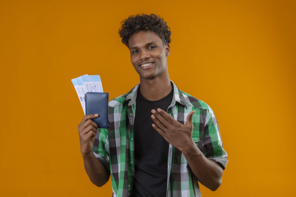 homem-jovem-viajante-afro-americano-segurando-passagens-aereas-sorrindo-alegremente-positivo-e-feliz-apresentando-se-com-o-braco-da-mao-olhando-para-camera-em-pe-sobre-fundo-laranja-1024x683 Como Planejar uma Mudança do Brasil para o Exterior em 2025: Passo a Passo Completo